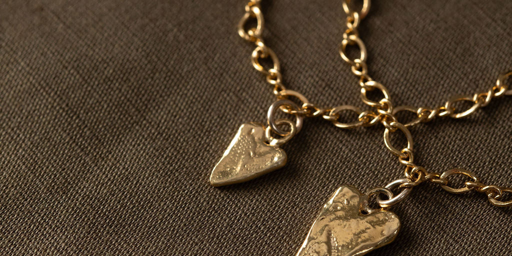 A close-up of a gold chain necklace with two textured arrowhead pendants resting on a dark brown fabric background.